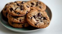 Fresh baked chocolate chip cookies stacked on green ceramic plate, showing gooey melted chips and golden brown edges, perfect for food and dessert blogs.