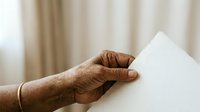 Senior male hand with wedding band holding blank white paper sheet against neutral beige curtain background for messaging or announcement purposes.