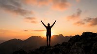Person celebrating success on mountain peak at sunrise with arms raised against dramatic orange sky and rocky landscape for motivation concepts.
