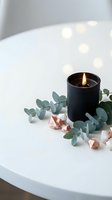 Black candle burning with eucalyptus leaves and rose gold crystals on white table creating cozy spa atmosphere with warm bokeh lights in background.