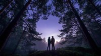 Silhouettes of two people standing together in a misty forest at dusk, surrounded by tall trees with purple twilight sky above, conveying connection and adventure.