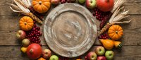 Autumn harvest arrangement with empty wooden plate surrounded by pumpkins, apples, berries and wheat on rustic wood table, overhead view for fall seasonal design.