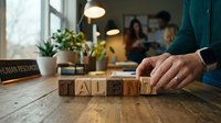Business professional arranging wooden letter blocks spelling TALENT on office desk with team meeting in background for human resources recruitment.