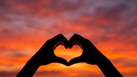 Silhouette of hands forming a heart shape against a vivid orange and red sunset sky, symbolizing love and romance.
