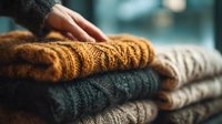 Hand touching stack of folded knitted sweaters in mustard yellow, dark green and beige colors, showcasing cozy winter clothing textures.