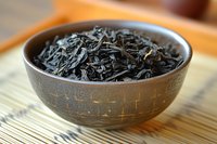 Dried black tea leaves in traditional ceramic bowl on bamboo mat, selective focus on loose leaf tea showing natural texture and organic quality for tea ceremony.