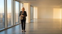 Professional businesswoman standing in modern office space with floor-to-ceiling windows overlooking city skyline during golden hour sunset.