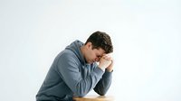 Young stressed businessman in gray shirt holding head in hands showing depression and mental health struggles isolated on white background.