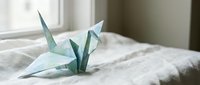 Delicate mint green origami paper cranes resting on soft white bedding in peaceful bedroom setting with natural window light creating serene minimalist atmosphere.