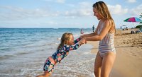 Young blonde mother in striped swimsuit playing with toddler daughter on sunny beach, holding hands and spinning together near ocean waves during summer vacation.