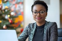 Professional African American woman with glasses and textured cardigan working on laptop during holiday season with decorated tree in background.