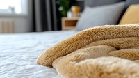 Soft plush beige blanket folded on white bedding creates cozy atmosphere in modern bedroom interior with natural morning light and blurred background.