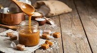 Homemade salted caramel sauce being poured from copper pot into glass jar with wooden spoon and caramel candies on rustic wooden table for dessert recipes.
