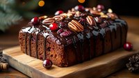 Chocolate loaf cake topped with dark chocolate ganache, fresh cranberries and pecans on wooden board. Festive holiday baking with rustic decorative elements.