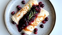 Delicate crepes filled and topped with fresh cranberry sauce and rosemary sprig served on gray ceramic plate, elegant breakfast or dessert presentation.