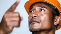 Asian male construction worker in orange hard hat looking up with determined expression while pointing upward, conveying leadership and vision in industrial setting.