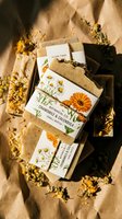 Handmade natural soap bars with chamomile and calendula botanical illustrations wrapped in kraft paper, displayed with dried flowers on neutral background for wellness and skincare branding.