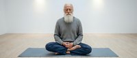 Senior man with long white beard and gray hair, wearing casual gray sweater and jeans, sitting in lotus position on blue yoga mat on wooden floor. Meditation and mindfulness practice.