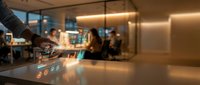 Barista pouring latte art into coffee cup at modern cafe counter with blurred customers and warm lighting in background.