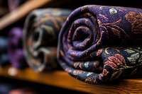 Rolled luxury fabric with intricate floral pattern in deep purple and copper tones, displayed on wooden shelf, showcasing elegant textile design and craftsmanship.