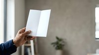 Mature male hand holding open blank white folded paper or brochure against neutral interior background with natural light, suitable for mockup, advertising, and marketing materials.
