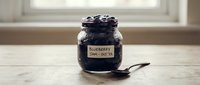 Homemade blueberry jam in glass jar with handwritten label and spoon on wooden surface near window for food photography and recipe content.
