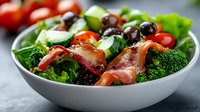 Fresh mixed salad with crispy bacon, broccoli, cherry tomatoes and black olives in white bowl. Healthy meal concept for restaurant menus and cooking blogs.