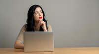 Thoughtful young Asian businesswoman with red lipstick working on laptop at wooden desk, hand on chin in contemplative pose for professional content.