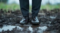 Business person wearing formal black leather shoes standing in wet mud and puddles, representing challenges and difficulties in professional journey.