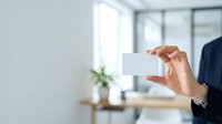 Mature professional man in business attire holding blank white business card in modern office environment with natural light and contemporary workspace background.