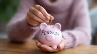 Person holding pink ceramic piggy bank with pension text written on it, inserting coin for retirement savings and financial planning on wooden table.