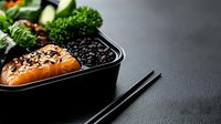 Fresh salmon fillet with black sesame seeds in meal prep container with black rice and green vegetables. Dark food photography with chopsticks.