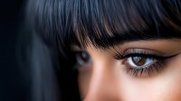 Close up portrait of young Asian woman with dramatic eye makeup, long eyelashes, and straight dark bangs against dark background. Professional beauty shot.