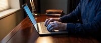 Professional businessman working on laptop computer at wooden desk in modern office environment with warm natural lighting from window.