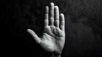 Open palm of a light-skinned hand raised against a dark textured background, conveying concepts of stop, refusal, protection, or communication in monochrome.