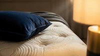 Close-up of navy blue and cream pillows arranged on a bed with wood grain texture, warm ambient lighting from bedside lamps creating cozy bedroom atmosphere.