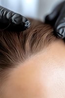 Professional hairstylist in black gloves applying hair dye to client's roots during coloring procedure at salon. Close-up view showing precise application technique.