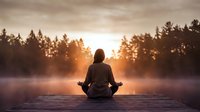 Woman meditating in lotus position on wooden dock at sunrise with misty lake and forest silhouette creating peaceful zen atmosphere for wellness and mindfulness.