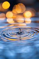 Water drop creating concentric ripples on blue water surface with golden bokeh lights reflecting in background, capturing moment of impact with crystal clarity.