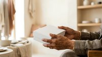 Senior man with dark skin tone holding white gift box in hands, wearing casual beige sweater indoors. Suitable for holiday, celebration, and lifestyle content.