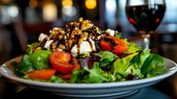 Fresh green salad topped with grilled chicken, cherry tomatoes, crumbled feta cheese and balsamic glaze on white plate, restaurant atmosphere with wine glass in background.