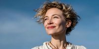 Young Caucasian woman with short curly blonde hair smiling joyfully against clear blue sky background, expressing happiness and freedom outdoors.