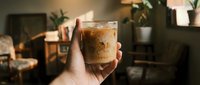 Hand holding a glass of iced coffee with creamy foam in a warm, modern interior setting with plants and ambient lighting.