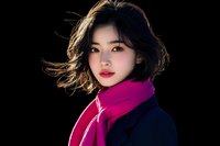 Young Asian woman with short dark hair wearing bright pink scarf and navy coat against black background, wind blowing through hair, dramatic lighting highlights facial features.