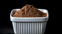 Ground coffee powder in white ceramic square ramekin on black background, dramatic lighting creates contrast between dark and light elements.