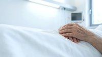 Elderly patient hands resting on white hospital bed sheets in modern medical facility room with natural lighting for healthcare and recovery concepts.