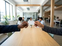Two diverse male professionals exchange business cards across wooden table in modern office with colleagues working in background, symbolizing networking and corporate partnership.