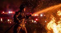 Firefighter in protective gear battles intense flames with water hose as sparks fly through smoky night air during emergency response operation.