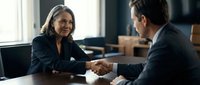 Professional businesswoman and businessman shaking hands across conference table in modern office setting during successful meeting or partnership agreement.