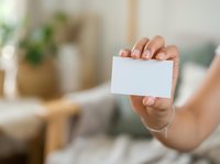 Woman holding blank white business card in modern home office environment with plants and natural lighting.